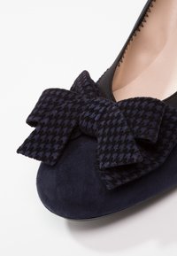 Chaussure plate en daim bleu marine avec un grand nœud en tissu pied-de-poule noir et bleu sur l'avant et une doublure intérieure beige.