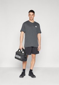Gray short-sleeve T-shirt with a small white logo, paired with black shorts and black shoes. Holding a black gym bag with a white logo.
