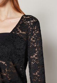 Femme portant un haut à manches longues en dentelle florale noire sur un sous-vêtement noir, montrant un motif transparent sur les manches et le haut de la poitrine.