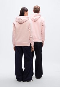 Ανοιχτό ροζ φούτερ με τσέπες καγκουρό και κορδόνια, συνδυασμένα με σκούρα τζιν φαρδιάς γραμμής. Το ύφασμα φαίνεται μαλακό με λεία υφή.