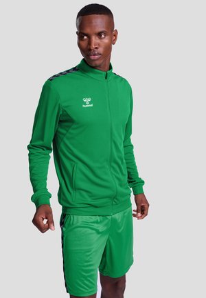 Groene sportieve jas en short set gemaakt van ademend materiaal. De jas heeft een ritssluiting aan de voorkant en contrasterende donkere strepen op de schouders.
