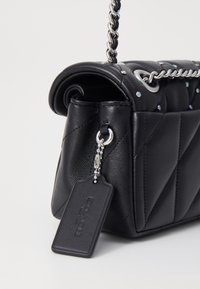 Svart kviltad läderhandväska med silverfärgad kedjestrap, små nitar och fäst rektangulär läderetikett på en bollar kedja.