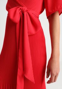 Vestido rojo con pliegues verticales, que presenta un cinturón de tela atado en un lazo. La textura suave y las mangas fluidas complementan el diseño.