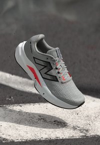 Baskets de sport grises avec une tige texturée, accents noirs et rouges, et une semelle intermédiaire blanche. Présente un logo bien visible sur le côté et les lacets.