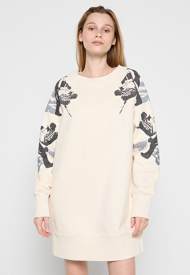 Junge Frau in einem cremefarbenen Langarmkleid mit schwarzen und grauen Skateboardgrafiken an Schultern und Ärmeln vor einem einfarbigen Hintergrund.