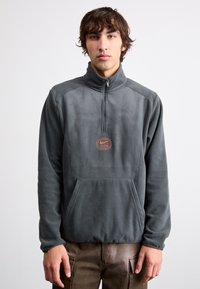 Γκρίζος φλις πουλόβερ με φερμουάρ μέχρι το μισό και μπροστινή τσέπη, με λογότυπο Nike Air σε πορτοκαλί. Μαλακή υφή και χαλαρή εφαρμογή.