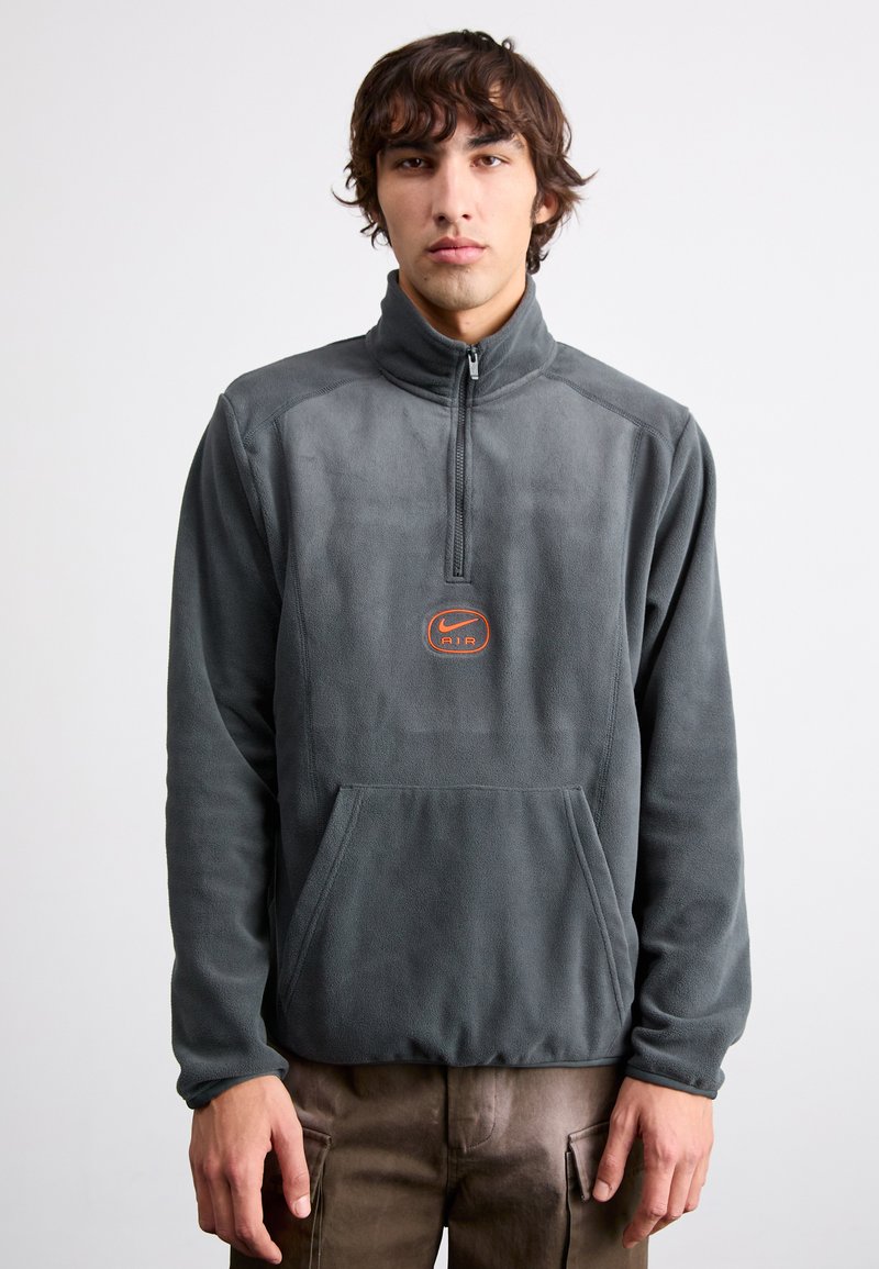 Γκρίζος φλις πουλόβερ με φερμουάρ μέχρι το μισό και μπροστινή τσέπη, με λογότυπο Nike Air σε πορτοκαλί. Μαλακή υφή και χαλαρή εφαρμογή.