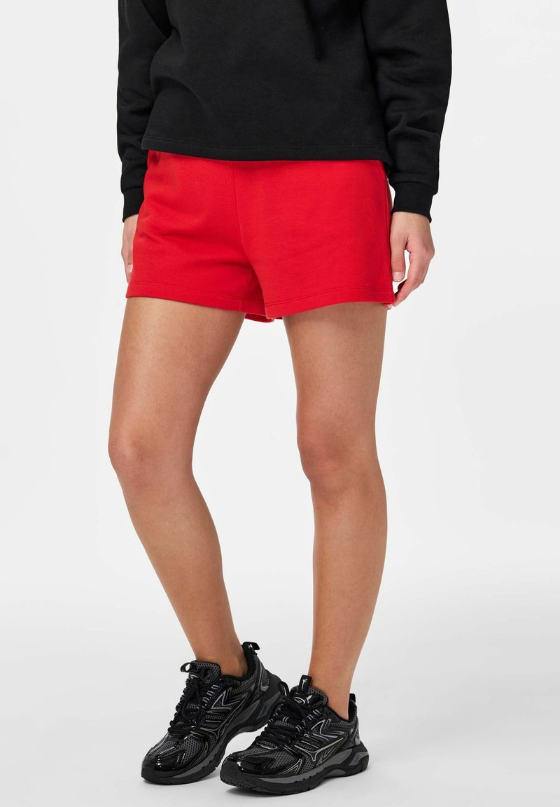 Personne portant un short rouge vif, un haut à manches longues noir et des baskets de sport noires, debout avec une jambe légèrement en avant.