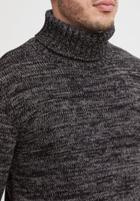 Svart og grå strikket genser med høy hals, med et strukturert mønster og tykk ribbestrikket krage. Har en tettsittende design uten fremtredende sømmer.