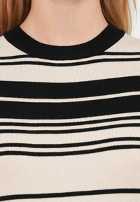 Gros plan sur une personne portant un pull en maille rayé horizontal beige et noir avec un col rond noir.