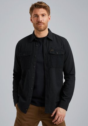 Black button-up shirt with a textured pattern, featuring two front pockets, a logo patch, and a classic collar, layered over a black t-shirt.