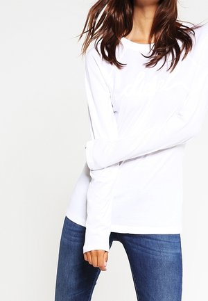 Vrouw draagt een eenvoudige witte lange mouw shirt en blauwe jeans, met één arm gebogen en hand nabij de heup tegen een effen achtergrond.