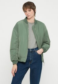 Zelena bomber jakna z gladko teksturo, rebrastim ovratnikom in zadrgo, oblečena čez sivo majico in visoko pasne jeans hlače.