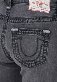I jeans in denim grigio presentano un design distintivo con cuciture nella tasca posteriore a forma di U. L'etichetta mostra il testo "World Tour" e un personaggio dei cartoni.