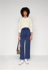 Ivory knitted cardigan with floral details, paired with high-waisted navy trousers featuring a bow tie, and white sneakers. Brown bag included.