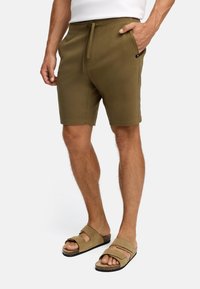 Homme portant un short olive avec cordon de serrage et des sandales beiges, debout avec une main dans la poche sur fond blanc.