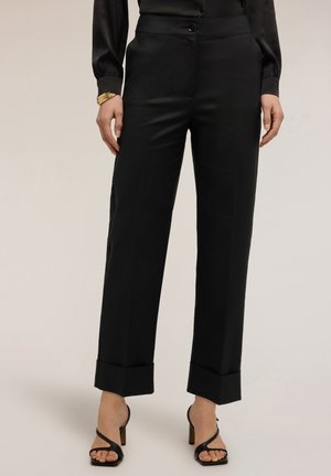 Kvinde iført sorte, højtaljede, skræddersyede bukser med opbuk ved benene, sort bluse med lange ærmer, guldbøjler og sorte hælede sandaler med stropper.