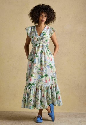 Kvinde med krøllet hår iført en hvid maxikjole med tropisk print og blå mokkasinsko, stående mod en beige baggrund.