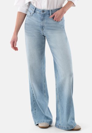 Jeans a gamba larga azzurro chiaro, indossati con scarpe bianche e una camicia bianca, una mano in tasca, in piedi davanti a uno sfondo semplice.