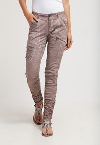 Jasnobrązowe spodnie cargo z wieloma kieszeniami, o dopasowanym kroju i marszczonej fakturze. Połączone z ozdobnymi, płaskimi sandałami.