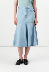 En person står iført et lys blått denimskjørt og en grønn topp, kombinert med svarte loafers, mot en nøytral bakgrunn.