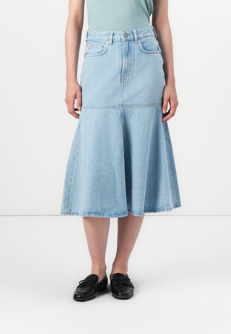 En person står iført et lys blått denimskjørt og en grønn topp, kombinert med svarte loafers, mot en nøytral bakgrunn.
