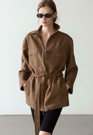 Frau trägt eine braune, mit Gürtel versehene Jacke über einem beigen Oberteil, schwarze Shorts und schwarze ovale Sonnenbrille, steht vor einem schlichten hellen Hintergrund.