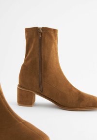 Botine din suede maro, cu vârf pătrat și toc bloc de înălțime medie. Prezintă un fermoar pe lateral și o textură netedă.