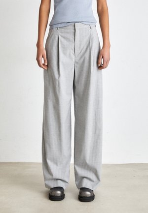 Person iført lysegrå pinstripe bukser med vide ben og metalliske sorte sko, stående på et neutralt gulv mod en hvid baggrund.