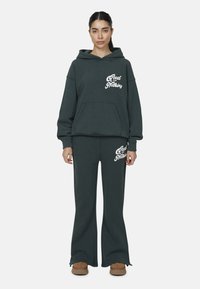 Kobieta w ciemnozielonym hoodie i odpowiadających szerokich spodniach, z białym napisem "Good for Nothing", stoi prosto na jednolitym tle.