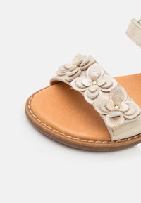 Sandales dorées légères avec une semelle plate ; dotées d'embellissements floraux superposés et d'une semelle intérieure en cuir naturel lisse.