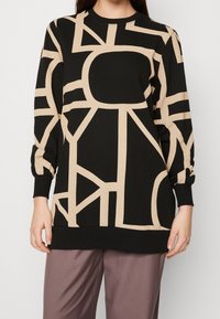 Kobieta ubrana w czarną bluzkę z długim rękawem, ozdobioną odważnymi, beżowymi, geometrycznymi liniami w stylu abstrakcyjnym, oraz luźne, brudnoróżowe spodnie, stojąca na jednolitym tle.