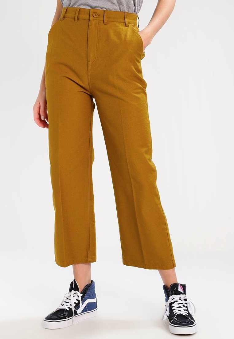 Pantalons larges jaune moutarde à taille haute, avec poches avant et fermeture à boutons, associés à des baskets noires et bleues.