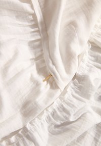 Tissu blanc texturé avec un drapé doux et des détails froncés. Présente une subtile étiquette en métal. Apparence légère et aérée.