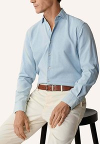 Muž sediaci na čiernom stolíku, oblečený v svetlomodrej košeli s gombíkmi, bielych nohaviciach a hnedom koženom opasku, s hlavou čiastočne otočenou do strany.