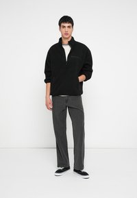 Mustast fliisist poollukuga pullover, mida on kombineeritud hallide joonitud pükstega. Modell seisab ühevärvilise valge tausta ees, jalas mustad tossud.