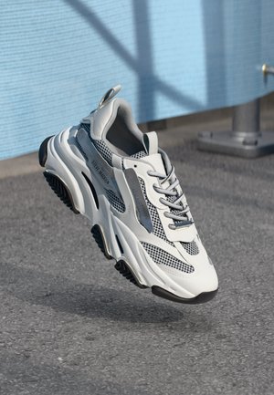 Scarpe da ginnastica bianche e grigie con tessuto in rete, colletto imbottito e suola in gomma spessa. Presentano una striscia riflettente e un design intricato.