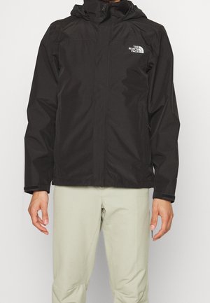Czarna kurtka z kapturem, z zamkiem z przodu i logo The North Face na piersi, noszona z jasnobeżowymi spodniami.