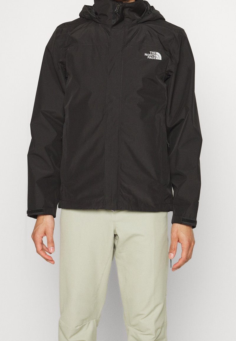 Veste noire à capuche avec fermeture éclair frontale et logo The North Face sur la poitrine, portée avec un pantalon beige clair.
