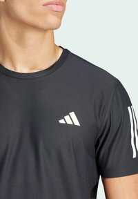 Maglietta atletica nera con tessuto strutturato. Presenta dettagli a tre strisce bianche sulla spalla e un logo bianco sul petto.