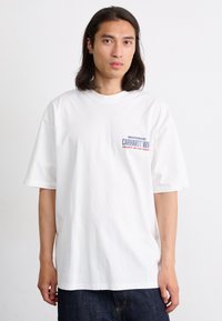 Hvit bomull t-skjorte med korte ermer, med en blå og rød "Carhartt WIP" logo på venstre bryst. Glatt tekstur, avslappet passform.