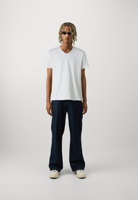 Tricou alb cu decolteu în V, cu o croială lejeră, asortat cu blugi largi din denim închisi la culoare. Accesoriile includ ochelari de soare subțiri și adidași de culoare deschisă.
