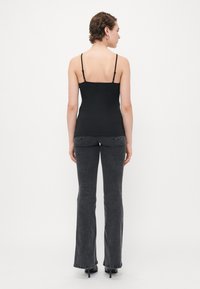 Femme aux cheveux courts et bouclés, portant un haut noir à bretelles fines, un jean bootcut noir délavé et des chaussures à talons noires, se tenant dos à la caméra.