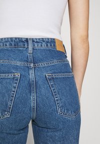 Džínové kalhoty s vysokým pasem v modré barvě s kontrastním prošíváním, dvěma zadními kapsami a hnědou koženou cedulkou na pase.