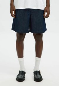Jeansshorts i mörkblått med rak skärning och upprullad fåll, kombinerat med vita strumpor och svarta loafers. Modellen står mot en enfärgad bakgrund.