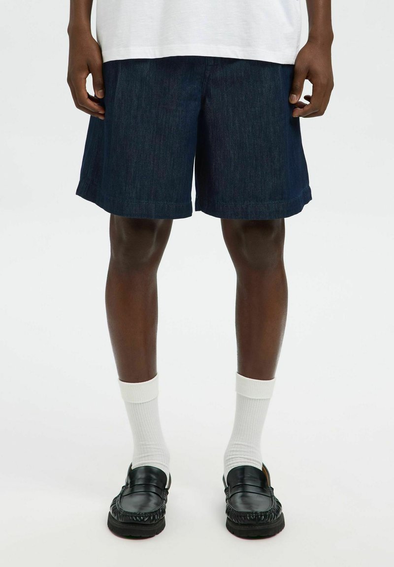 Jeansshorts i mörkblått med rak skärning och upprullad fåll, kombinerat med vita strumpor och svarta loafers. Modellen står mot en enfärgad bakgrund.