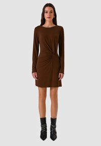 Abito marrone a maniche lunghe con dettaglio arricciato sul davanti, scollo rotondo e tessuto morbido, abbinato a stivaletti neri con dettagli borchiati.