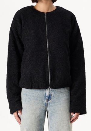 Person iført en sort tekstureret jakke med lynlås og lange ærmer samt lyseblå jeans med høj talje mod en ensfarvet baggrund.