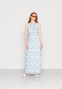 Vestido maxi floral com decote halter, base branca com flores azuis e pêssego. Combinado com sapatos brancos e óculos de sol laranja.