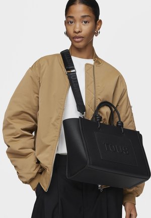 Black leather tote bag with embossed "TOUS" logo, featuring two short handles and a detachable strap, paired with a beige bomber jacket.